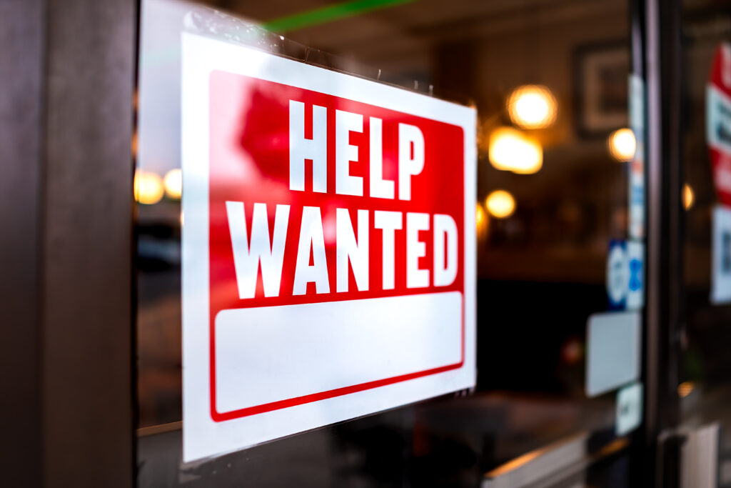 Sign text closeup for help wanted with red and white colors by entrance to store--employment rates are causing wage rates to grow.